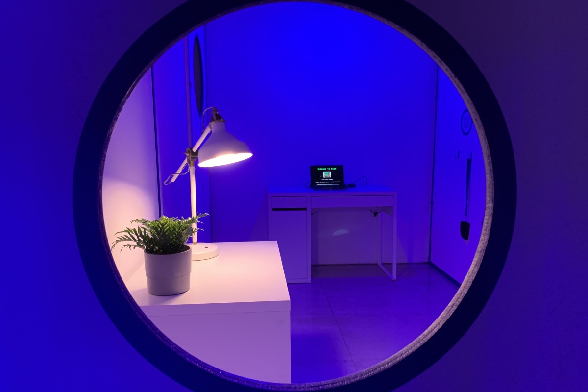 A round window looking into a room bathed in purple light with a white desk, lamp and plant on top.